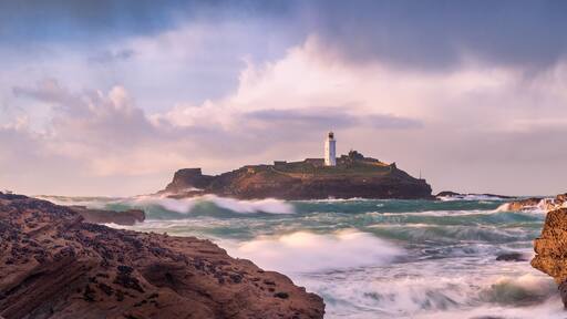 A rocky coastline with a lighthouse on a small island, surrounded by turbulent sea waves.