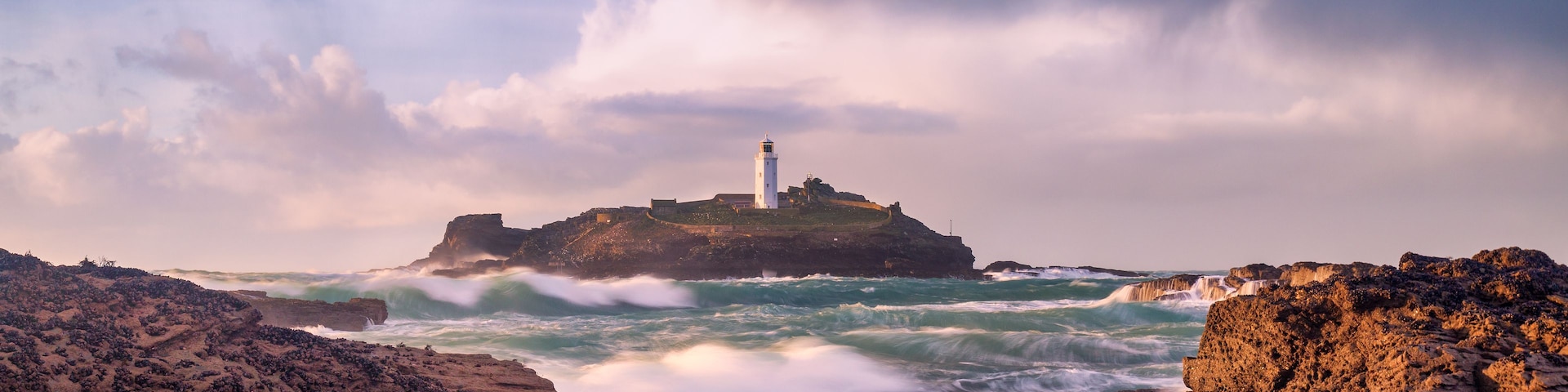 Un litoral rocoso con un faro en una pequeña isla, rodeado por olas marinas agitadas.