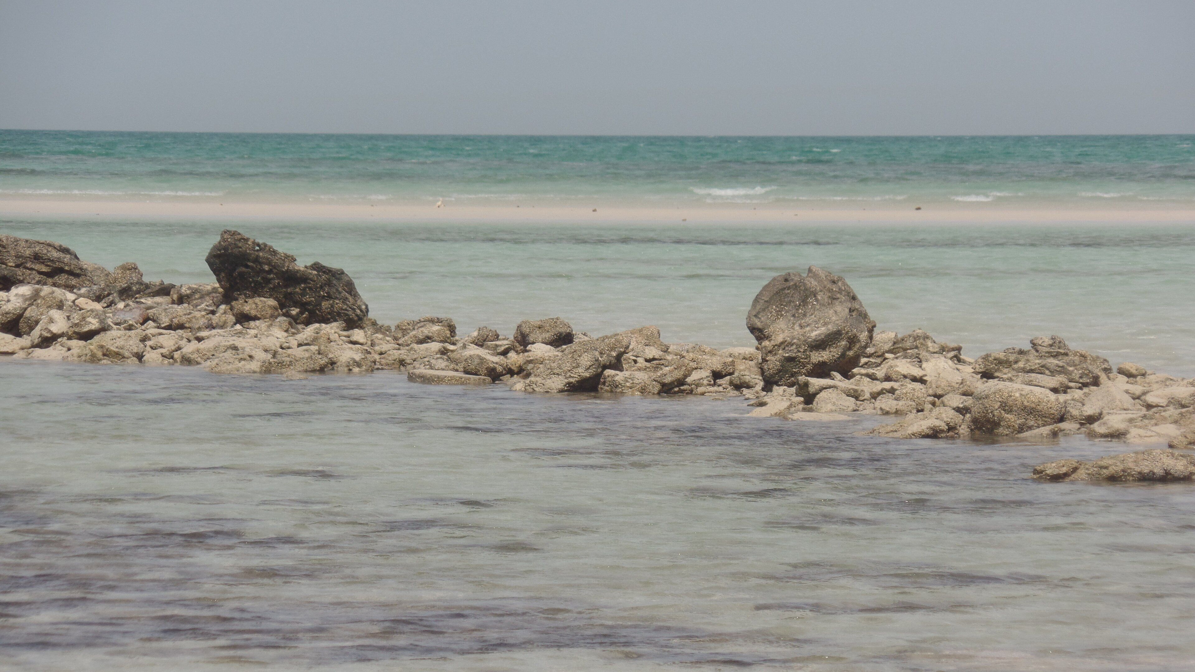 A rocky shoreline with clear turquoise water and a sandy beach in the background.