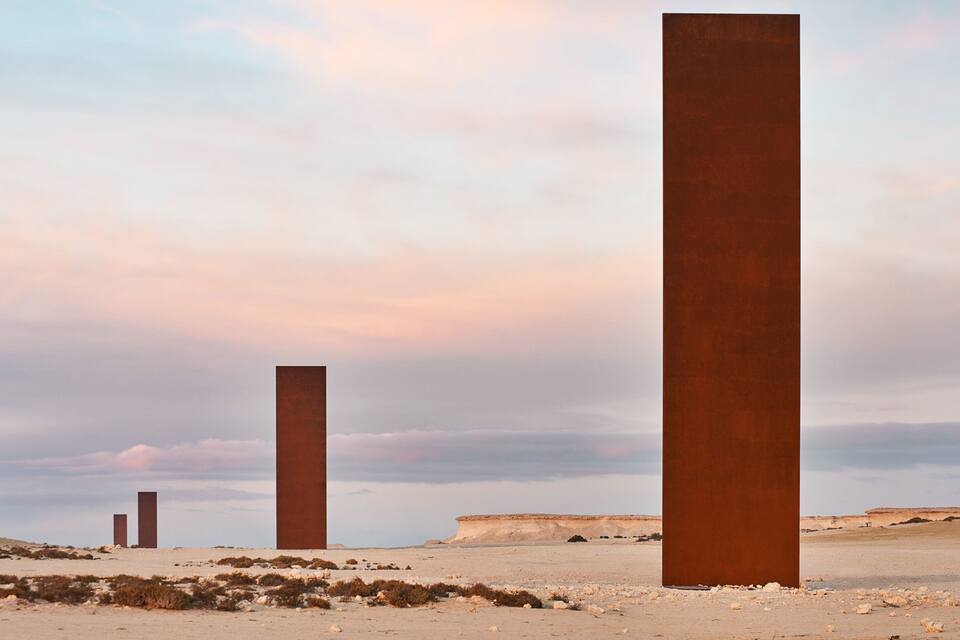 Three tall, rust-colored metal structures in a desert landscape with a cloudy sky.