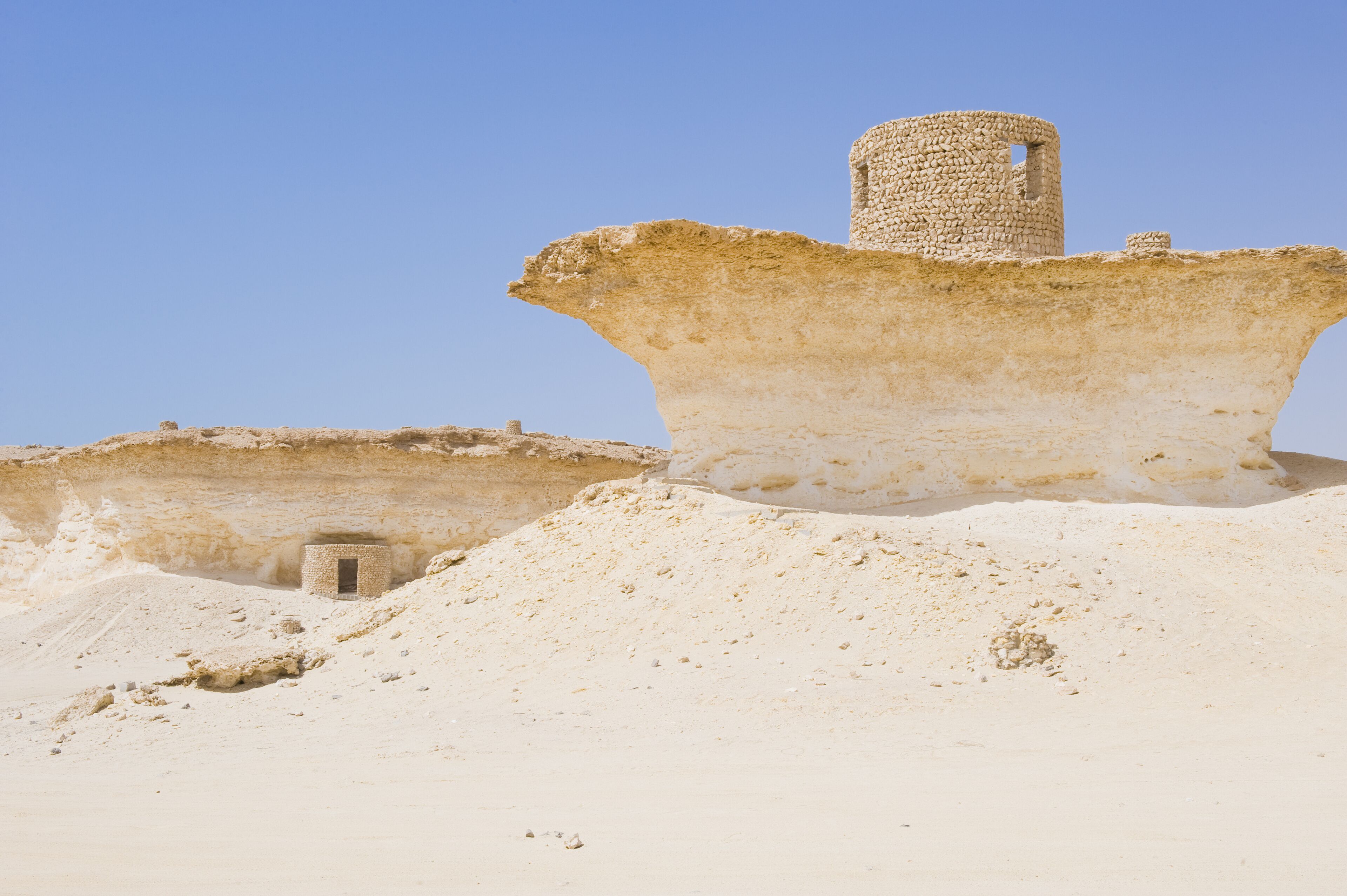 Paisaje desértico con ruinas antiguas, incluyendo una gran estructura de piedra y un edificio más pequeño.