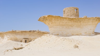 Paisagem desértica com ruínas antigas, incluindo uma grande estrutura de pedra e um edifício menor.