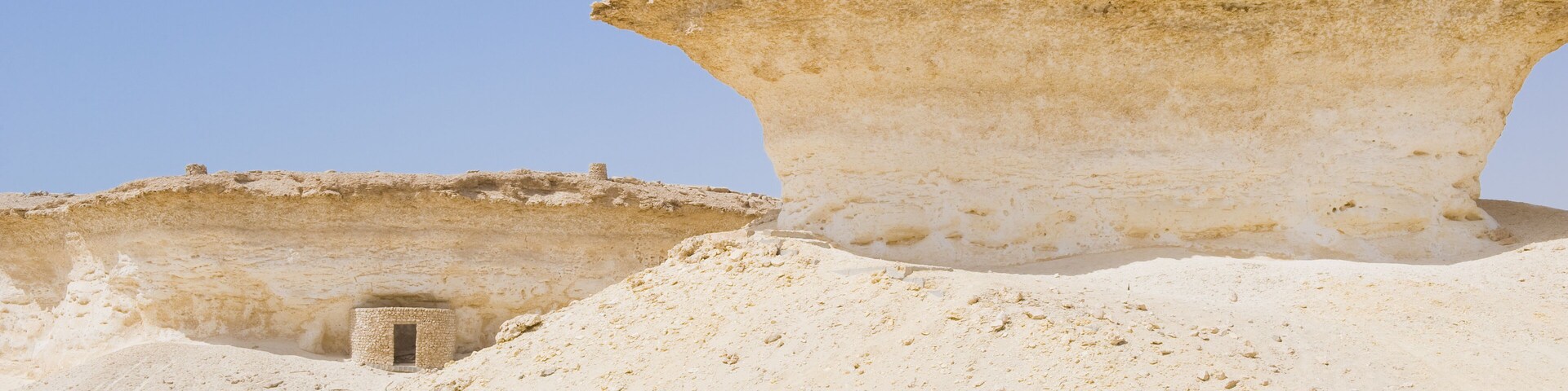 Paisaje desértico con ruinas antiguas, incluyendo una gran estructura de piedra y un edificio más pequeño.