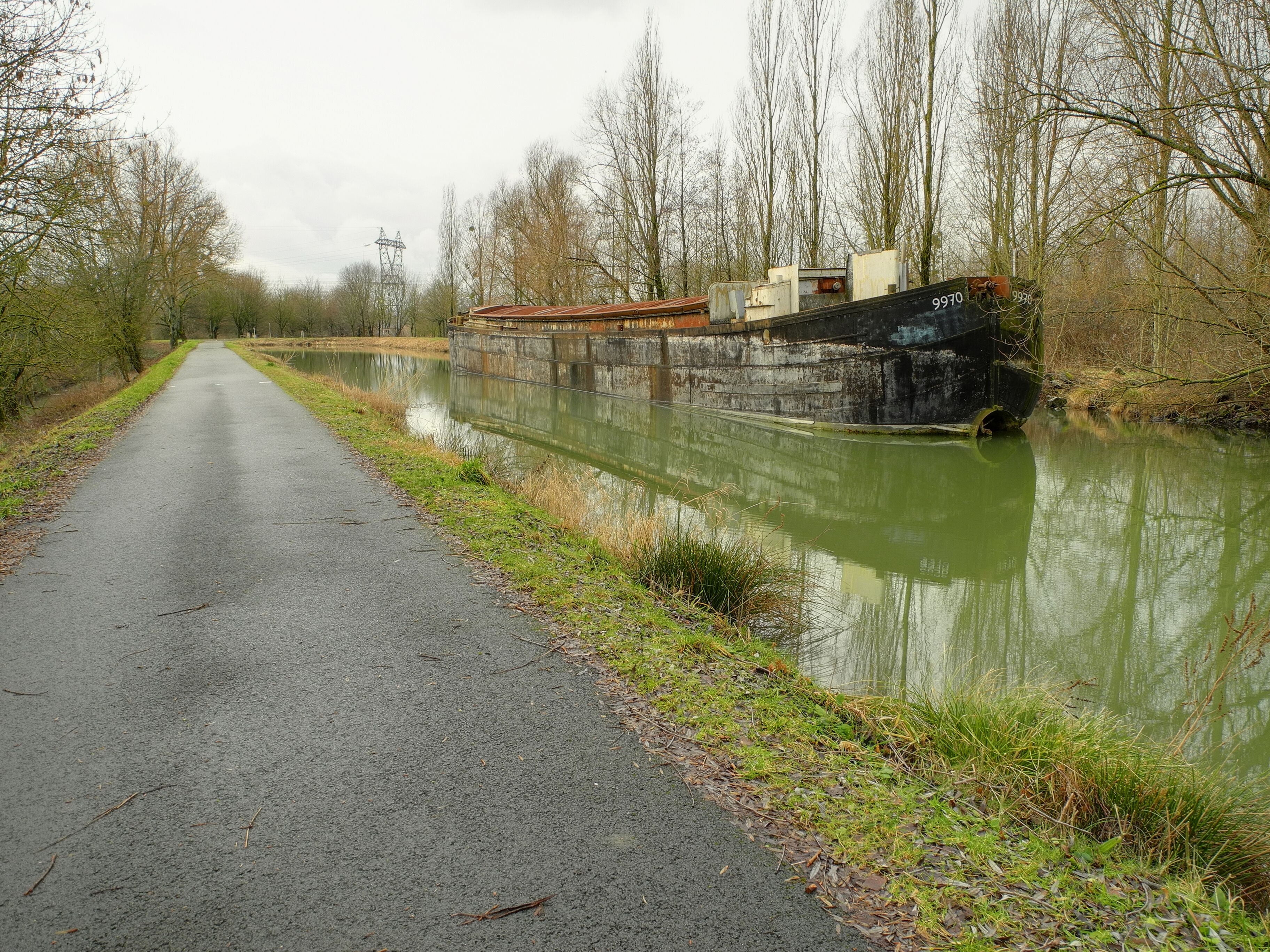 A narrow road beside a canal with a rusted boat and a power line tower in the background.