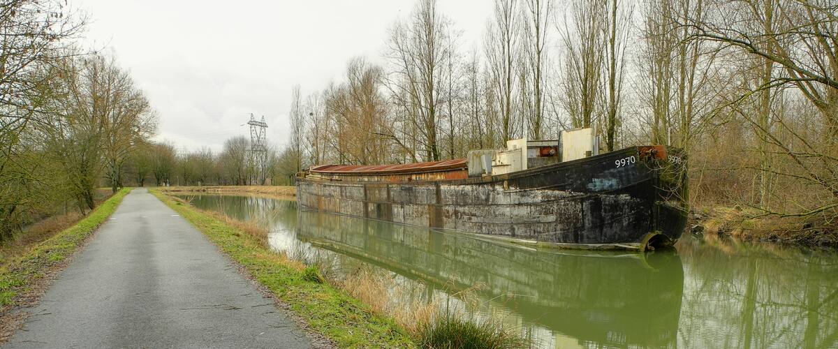 A narrow road beside a canal with a rusted boat and a power line tower in the background.