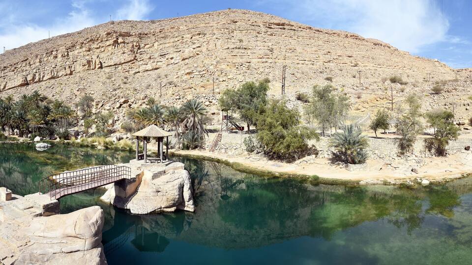 A clear mountain lake with a bridge, palm trees, and a small pavilion.