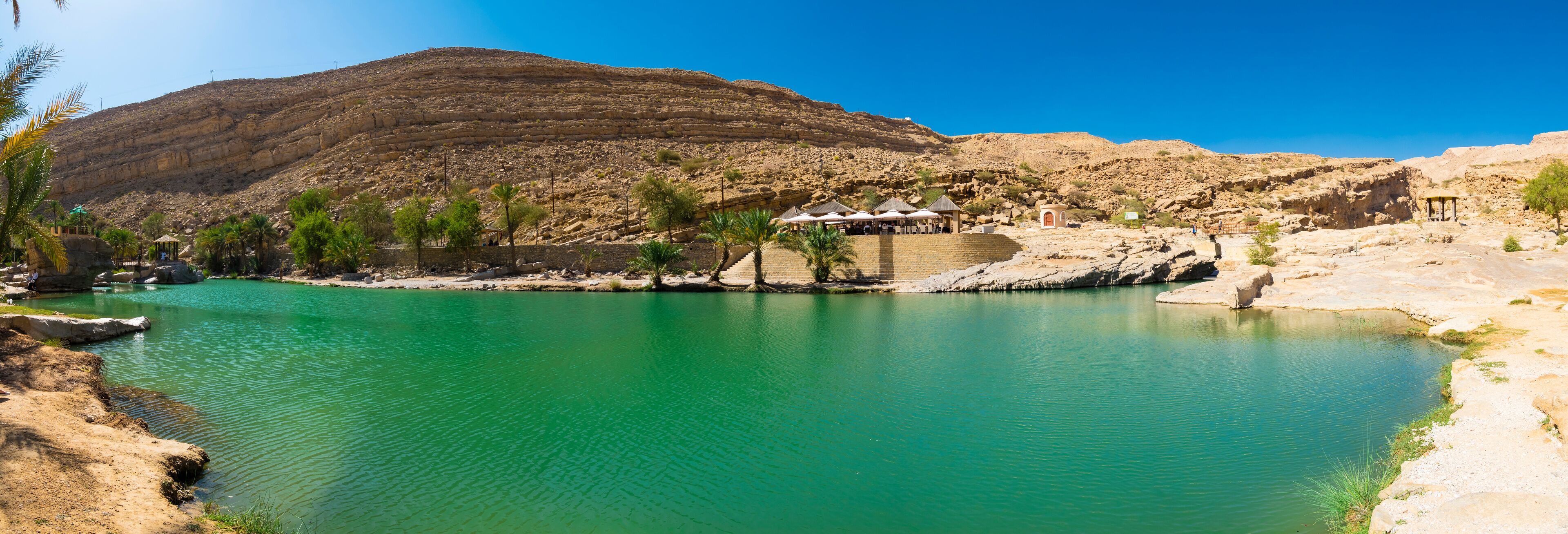 A turquoise lake surrounded by rocky terrain and palm trees, with a mountain in the background and a small pavilion by the water's edge.