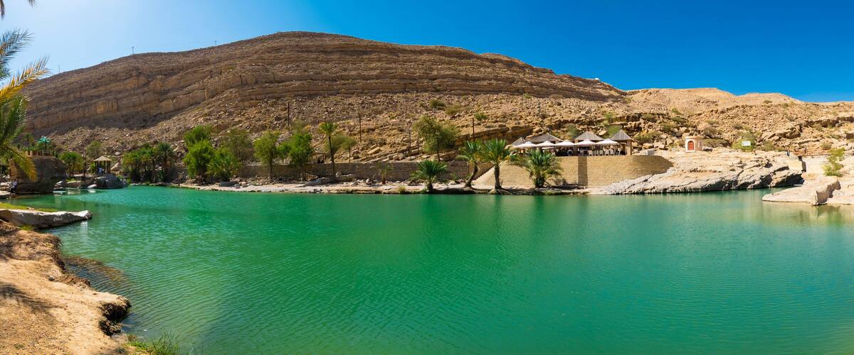 A turquoise lake surrounded by rocky terrain and palm trees, with a mountain in the background and a small pavilion by the water's edge.