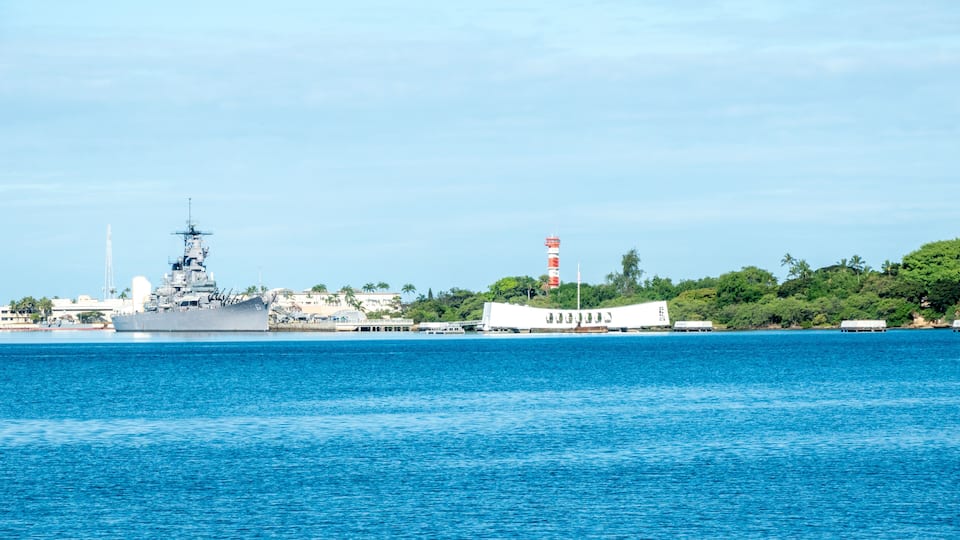 A large naval ship docked at a port with a lighthouse and lush greenery in the background.
