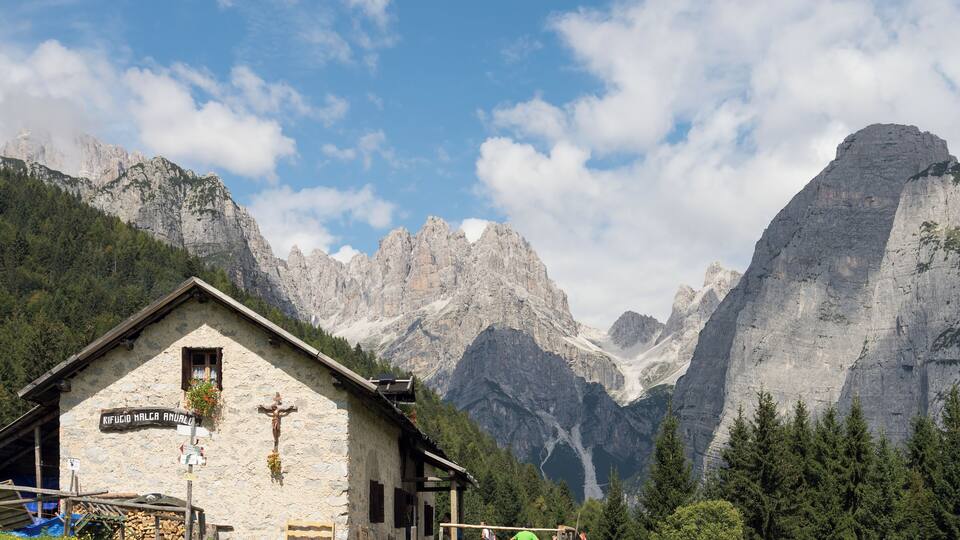 A stone building with a cross, surrounded by mountains and trees.
