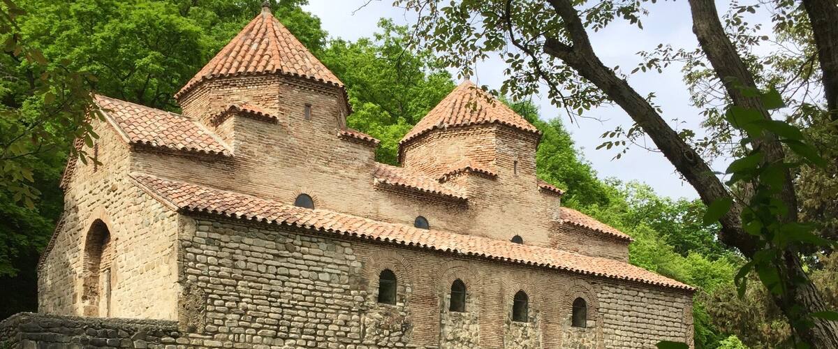 A historic stone building with a tiled roof, surrounded by lush greenery.
