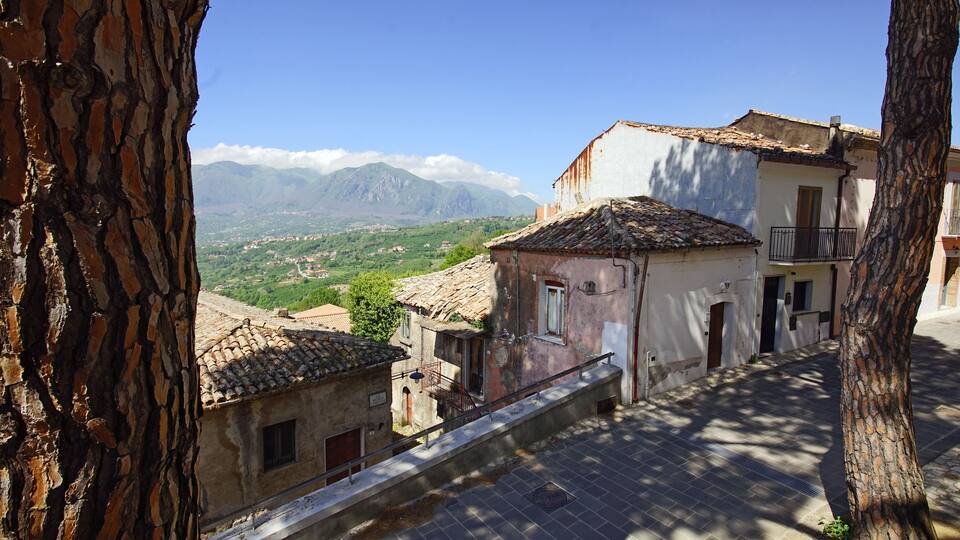 A narrow street with traditional buildings, a tree, and a mountainous landscape in the background.