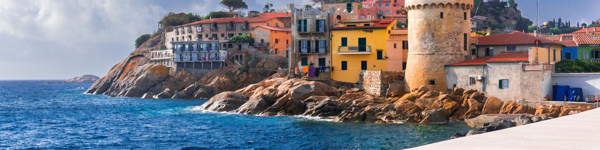 Perfect tiny seaside village of "Giglio Porto" with multi colored houses, an ancient defensive tower and a rocky coastline against a deep blue Mediterranean sea. - Giglio Island, Tuscany, Italy