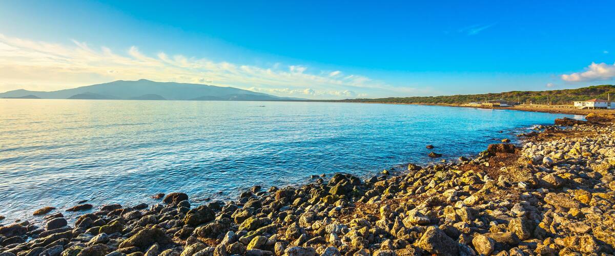 A rocky shoreline with clear blue water, a distant mountain, and a few buildings on the right.