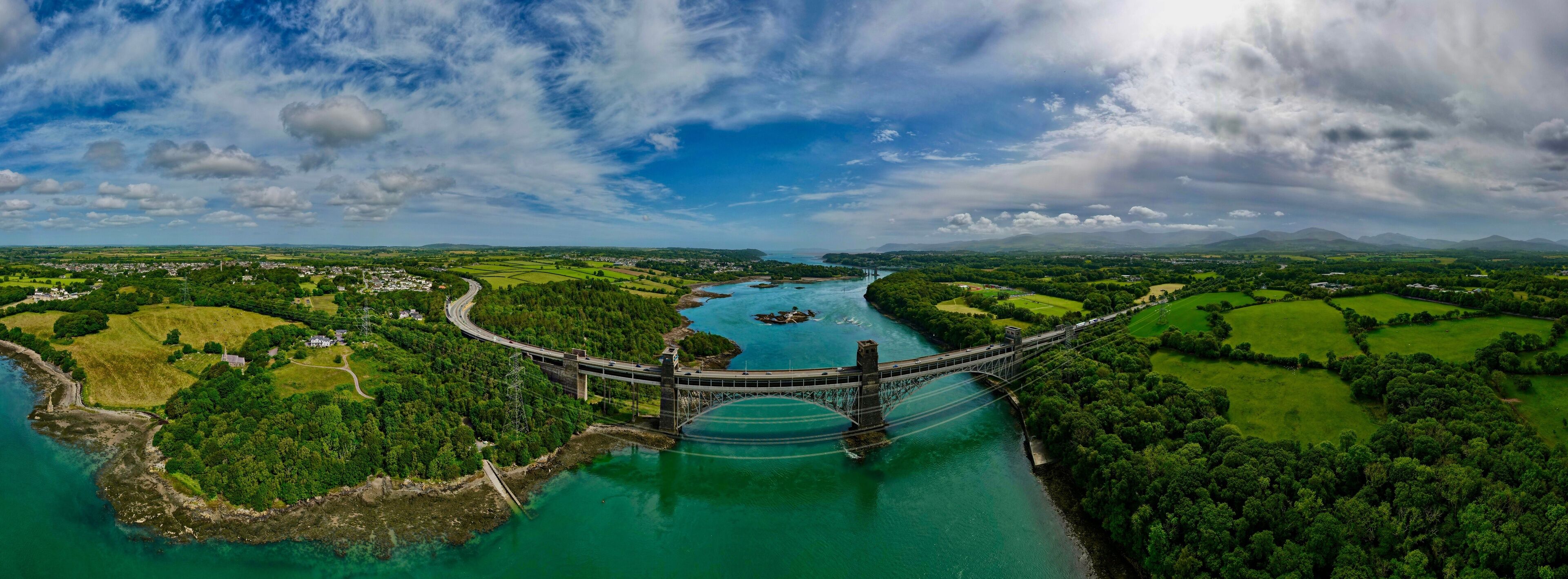 A suspension bridge over a river with surrounding green fields and mountains in the background.