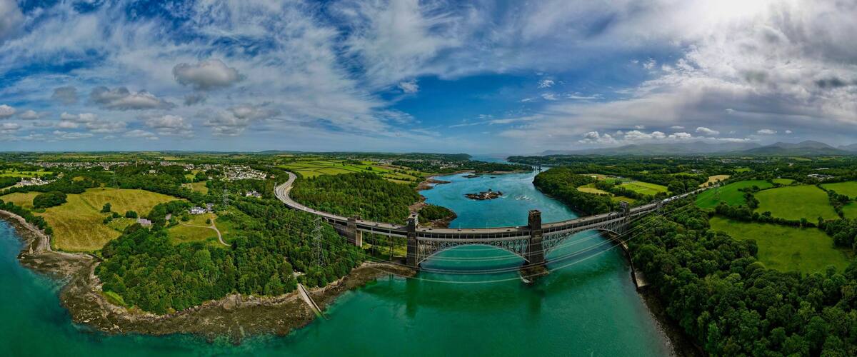 A suspension bridge over a river with surrounding green fields and mountains in the background.