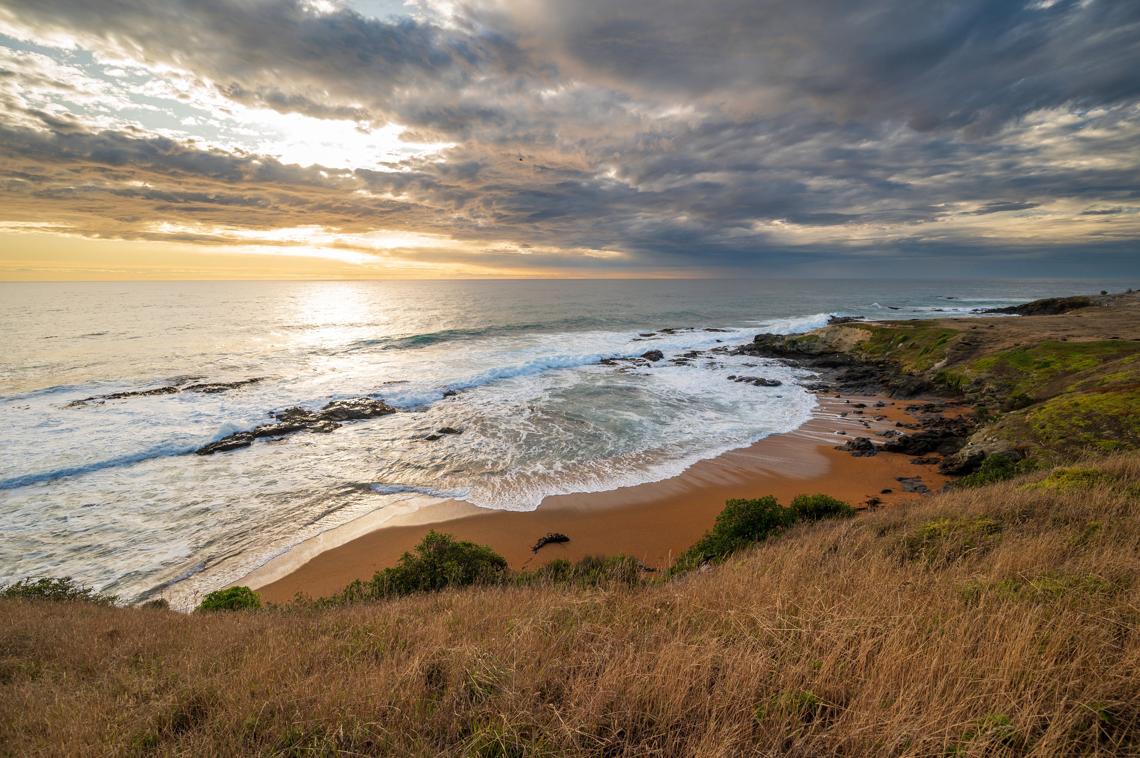 A coastal landscape with a sandy beach, rocky shoreline, and a grassy cliff.