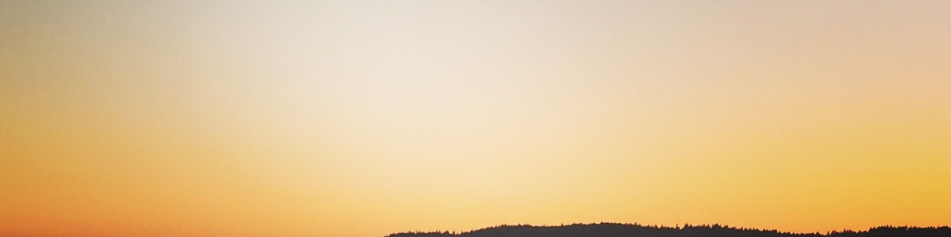 Ein ruhiger See bei Sonnenuntergang mit mehreren Booten auf dem Wasser und einem bewaldeten Hügel im Hintergrund.