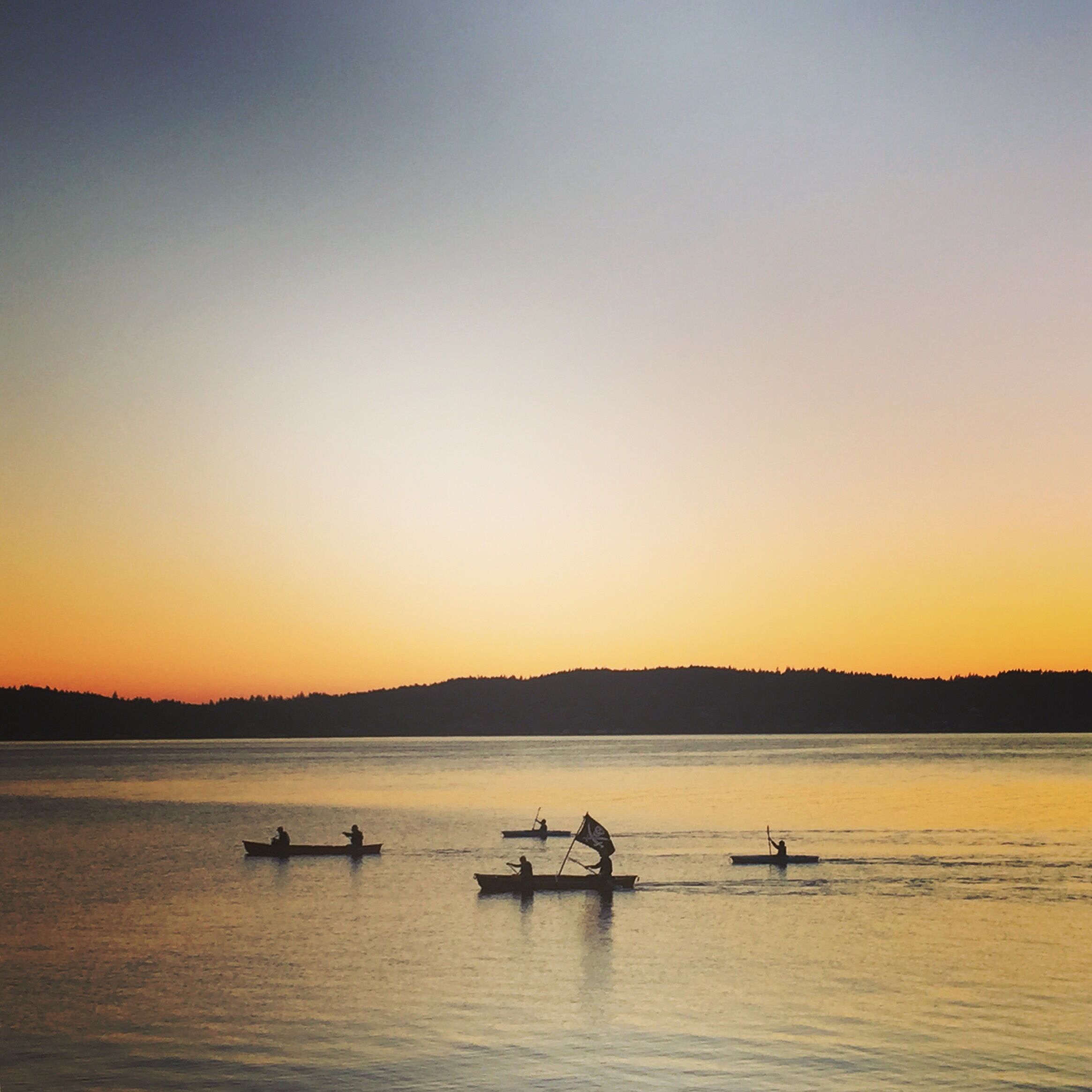 A calm lake at sunset with several boats on the water and a forested hill in the background.