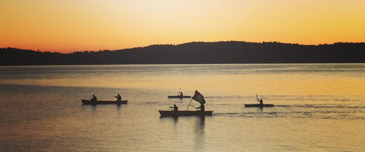 A calm lake at sunset with several boats on the water and a forested hill in the background.