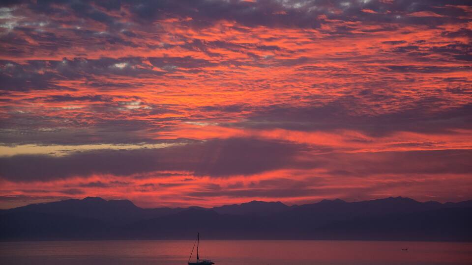 A sunset over a calm lake with a sailboat, mountains in the background, and a silhouette of trees in the foreground.