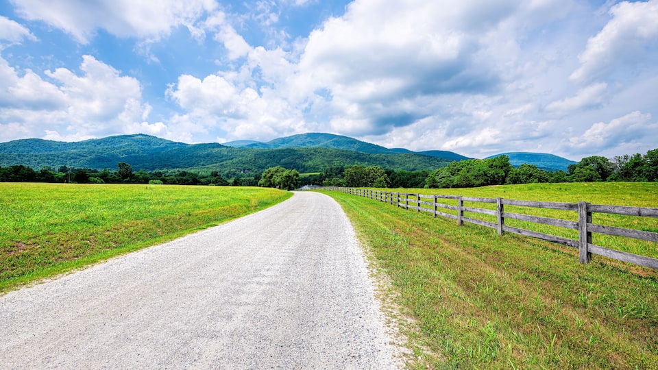 A gravel road curving through a green field with a wooden fence on one side and mountains in the background.