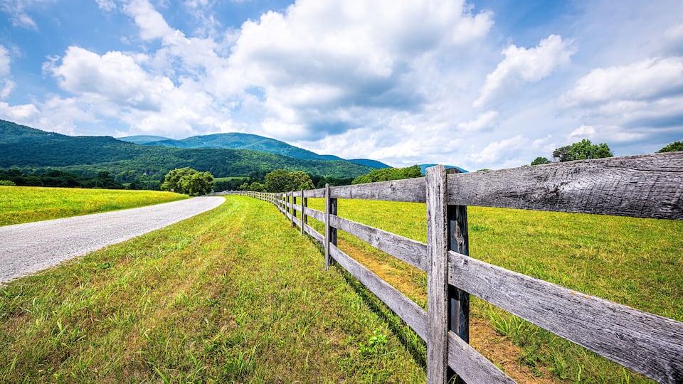 A rural landscape with a gravel road, wooden fence, green fields, and mountains in the background.
