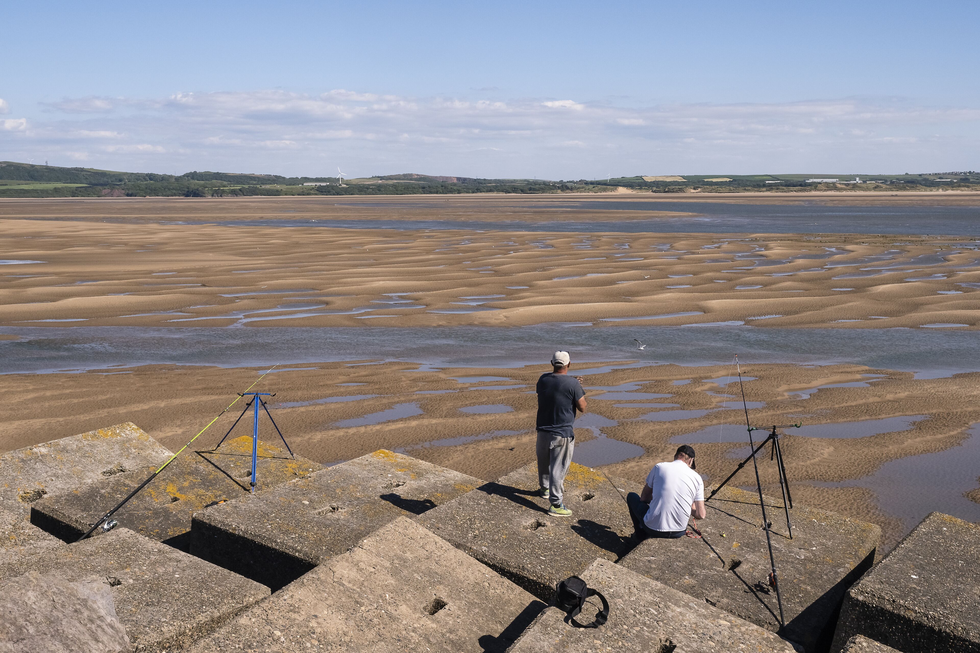 A coastal landscape with a wide expanse of wet sand and water, two fishing tripods, and a distant wind turbine.