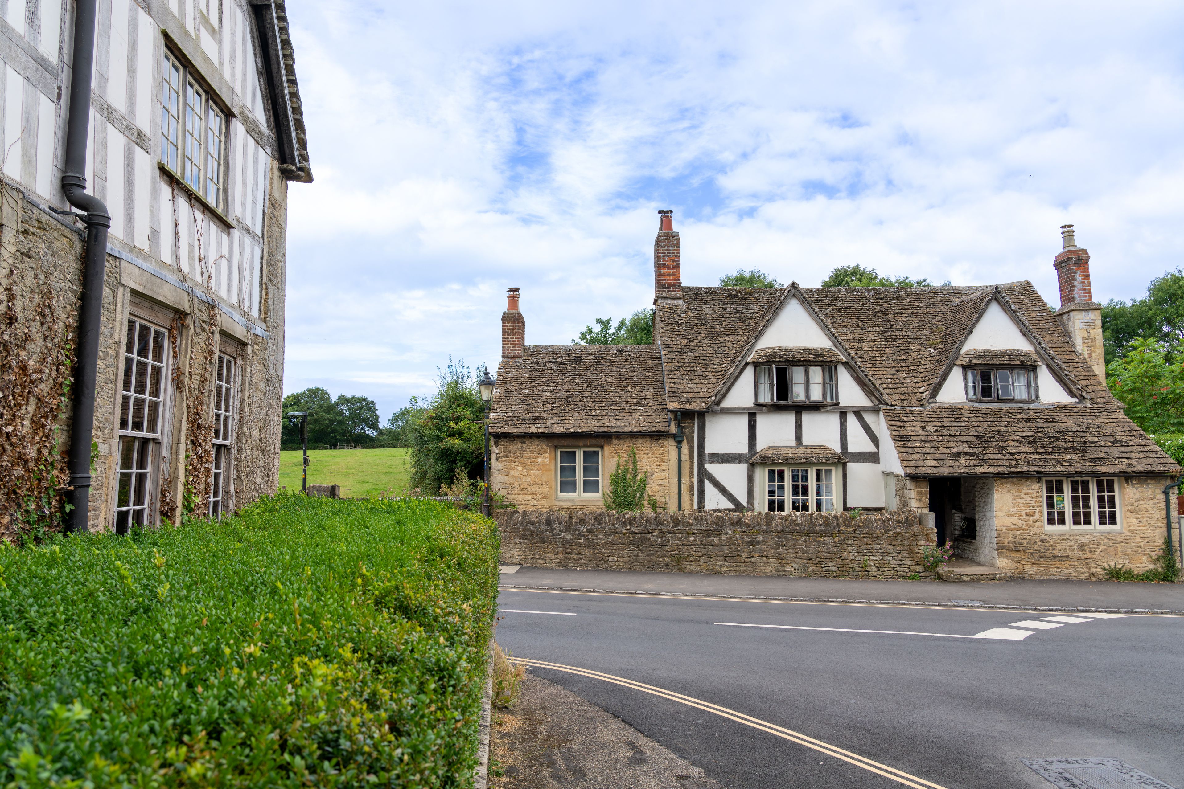 A traditional half-timbered house with a stone chimney, a paved road, and a hedge in the foreground.