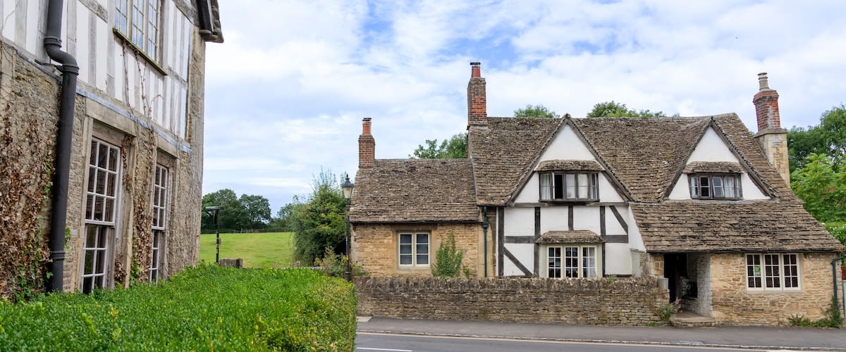 A traditional half-timbered house with a stone chimney, a paved road, and a hedge in the foreground.