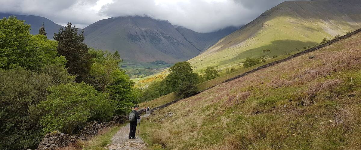 A hiker on a stone path in a mountainous area with lush greenery and a cloudy sky.