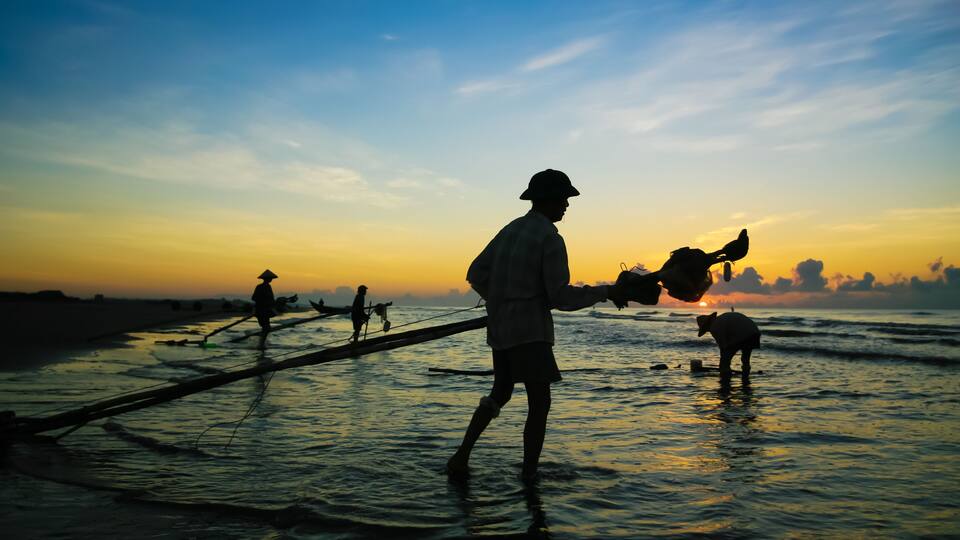 A person in traditional attire working with fishing nets on a beach at sunset.