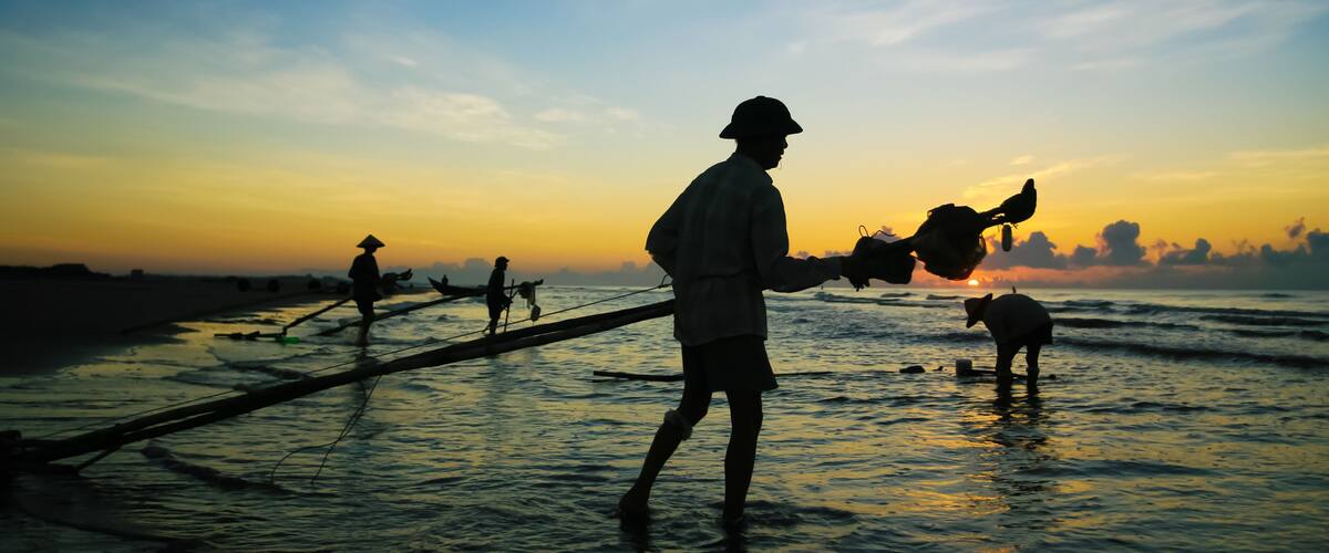 A person in traditional attire working with fishing nets on a beach at sunset.