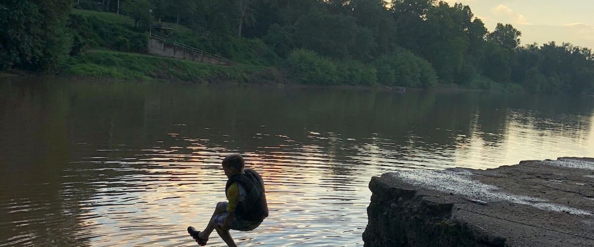 A person sitting on a concrete ledge by a calm river, surrounded by greenery and trees.