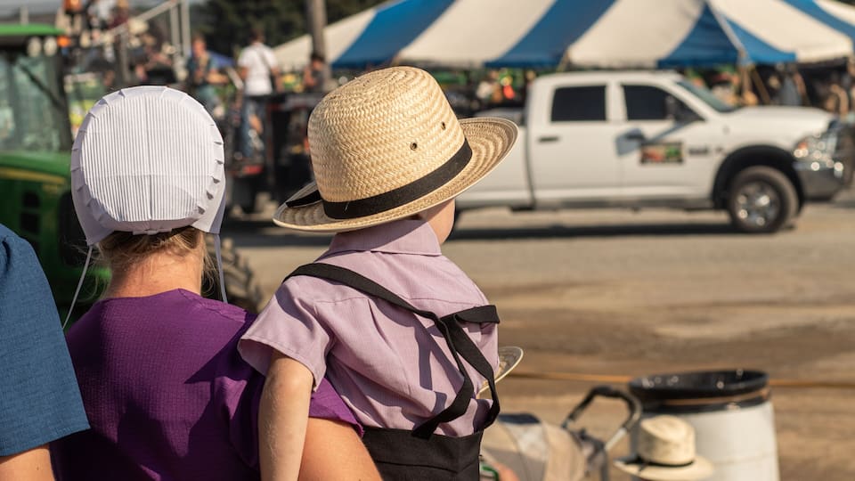 A person wearing a straw hat and a purple shirt with suspenders, standing in a parking lot with a white pickup truck and tents in the background.