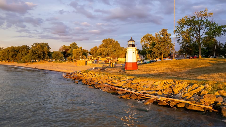 A lighthouse by a rocky shoreline with a beach and trees in the background.