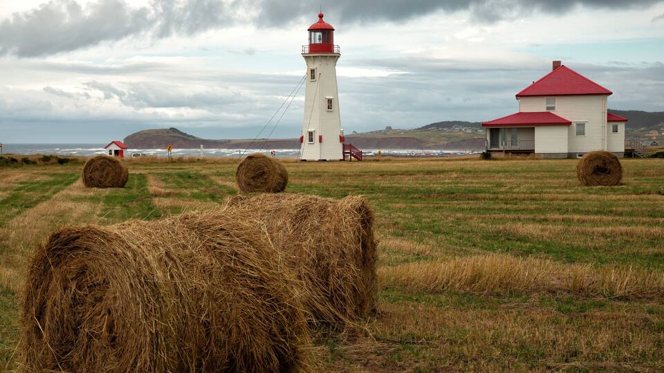 A lighthouse, a red-roofed house, and hay bales in a grassy field with the ocean in the background.