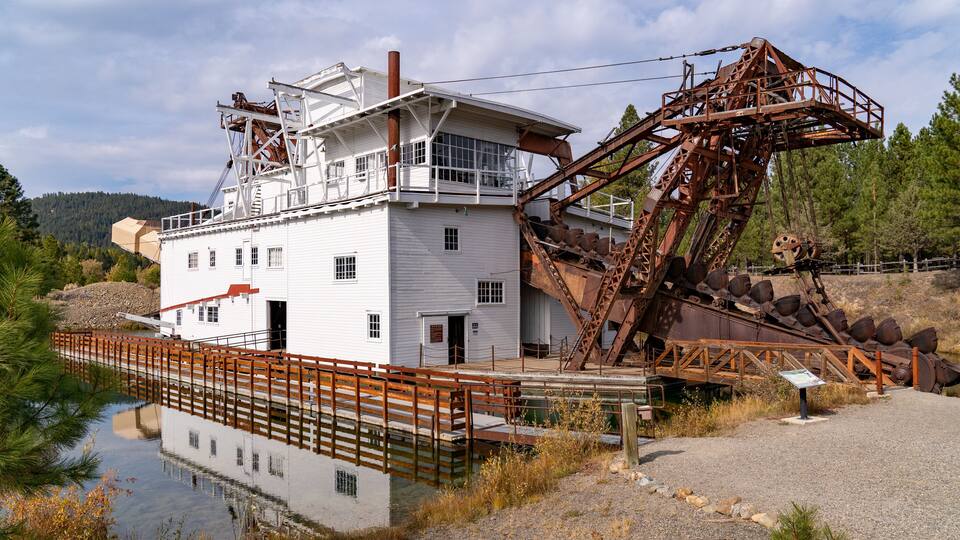 A white industrial building with a large metal structure, a wooden bridge over a body of water, and a forested hillside in the background.