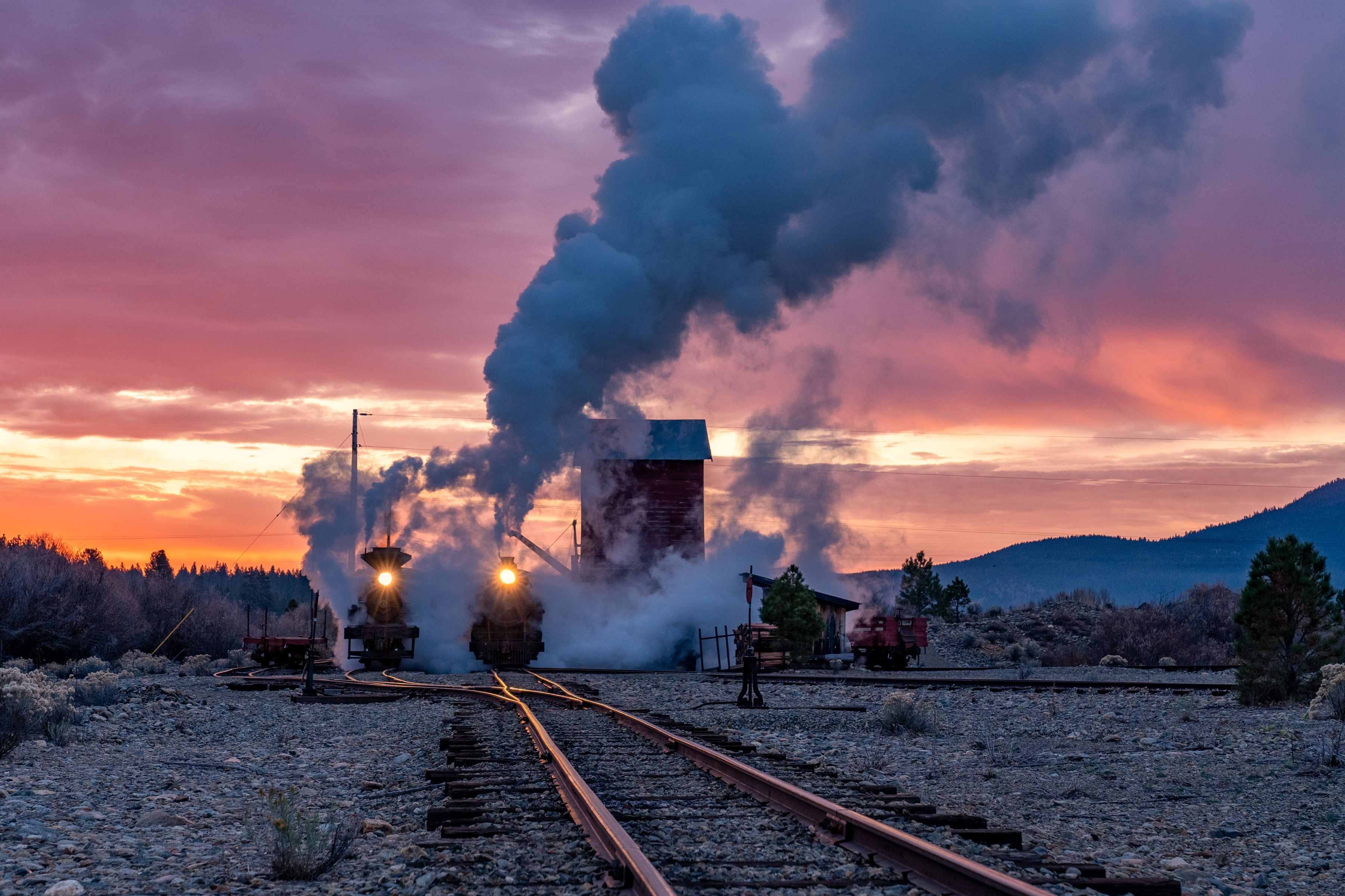 Una locomotora de vapor emitiendo humo, vías de tren y un paisaje montañoso al atardecer.