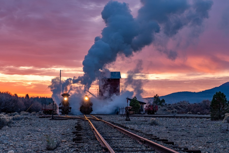 Una locomotora de vapor emitiendo humo, vías de tren y un paisaje montañoso al atardecer.