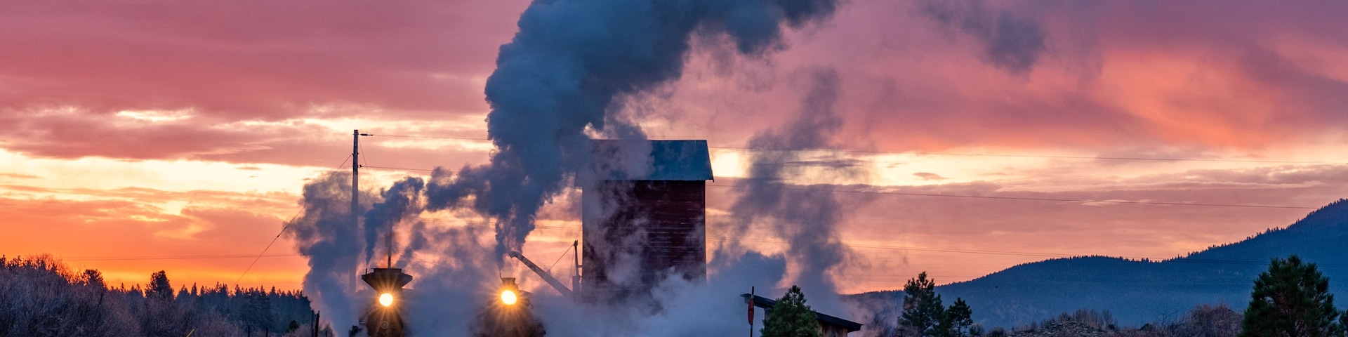 Una locomotora de vapor emitiendo humo, vías de tren y un paisaje montañoso al atardecer.