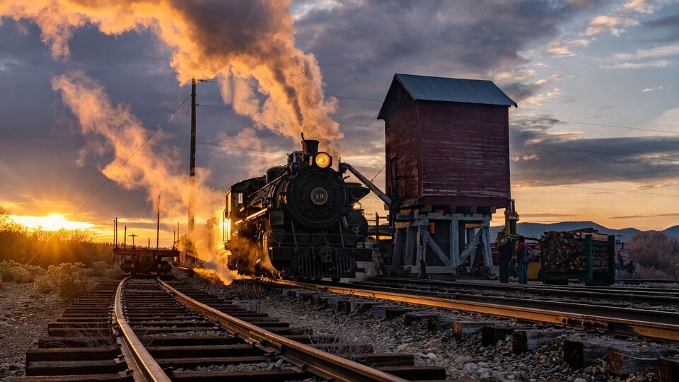 A steam locomotive emitting smoke, a wooden building, and railway tracks during sunset.
