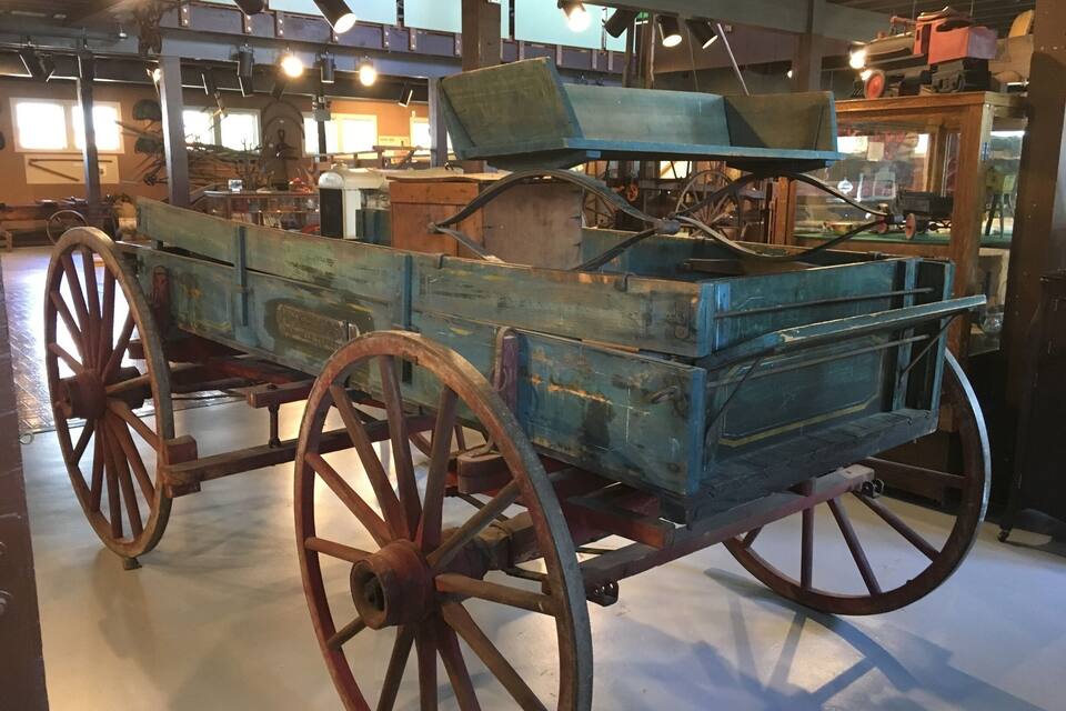 An antique wooden wagon with metal wheels and a flatbed, displayed indoors with various lighting fixtures and other historical items in the background.