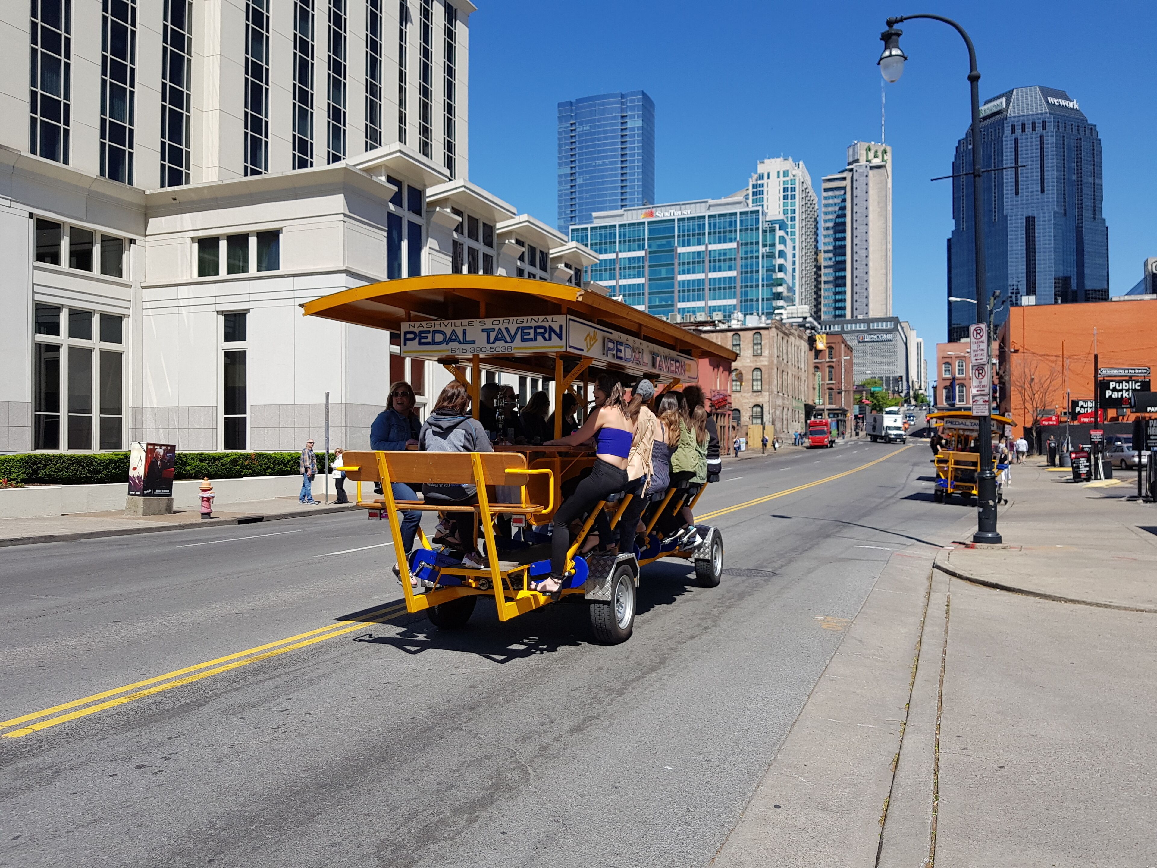 Un tranvía amarillo con pasajeros, con la leyenda "Nashville's Original Pedal Tavern", en una calle de la ciudad con edificios altos al fondo.