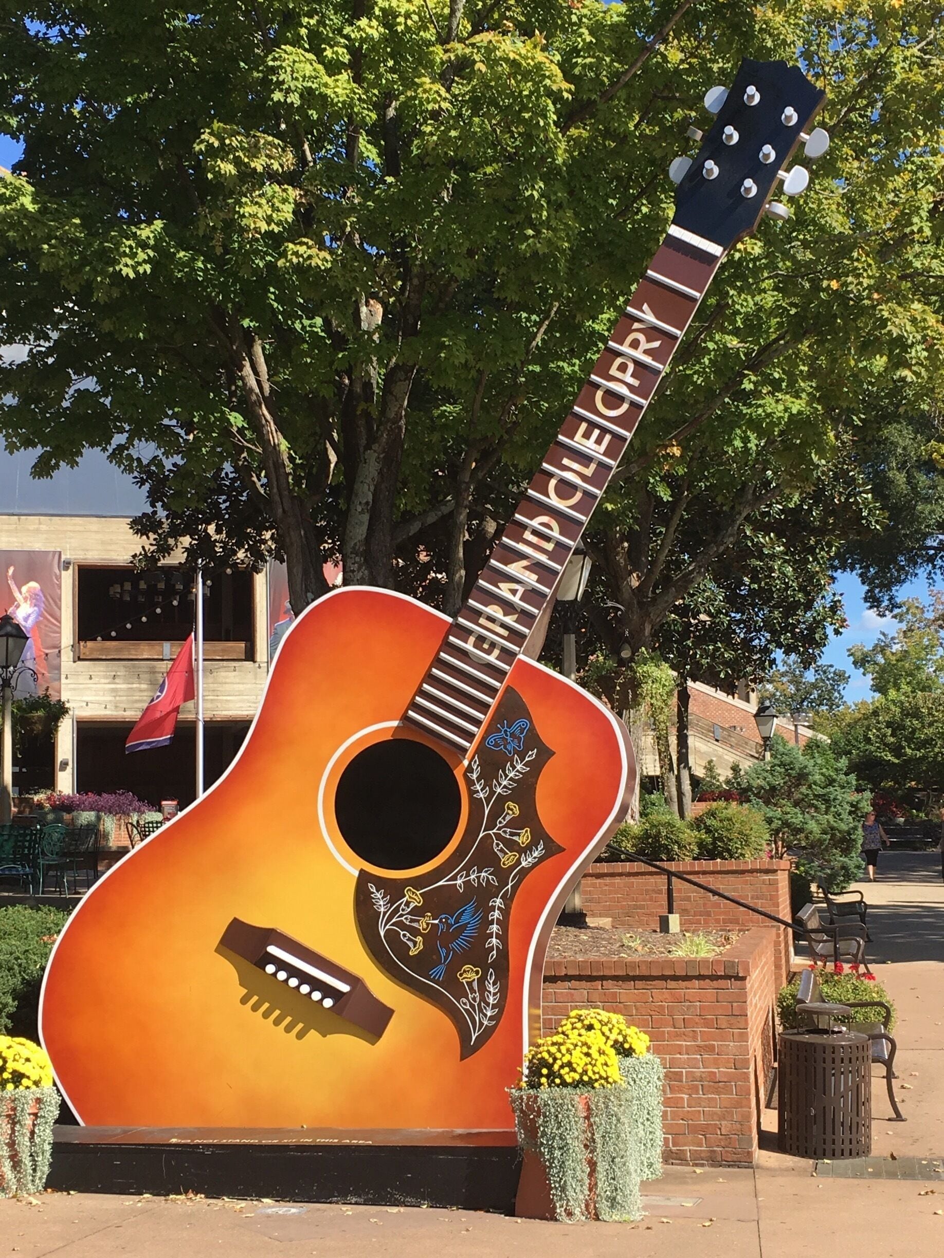 Eine große Gitarrenskulptur mit roten und braunen Farbverläufen, umgeben von Grünpflanzen und einer Ziegelwand.