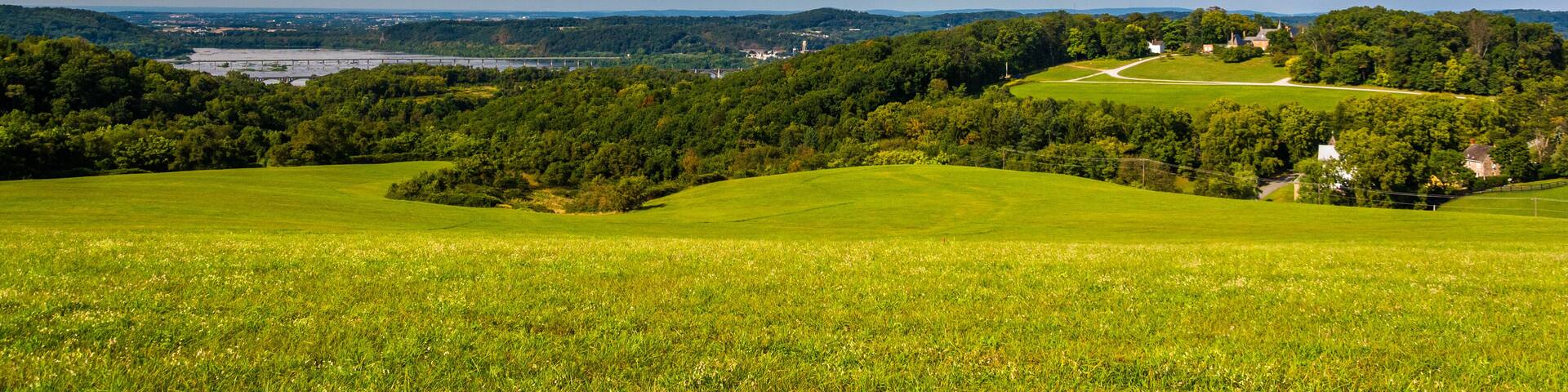 A green landscape with rolling hills, a river, and a bridge in the distance.