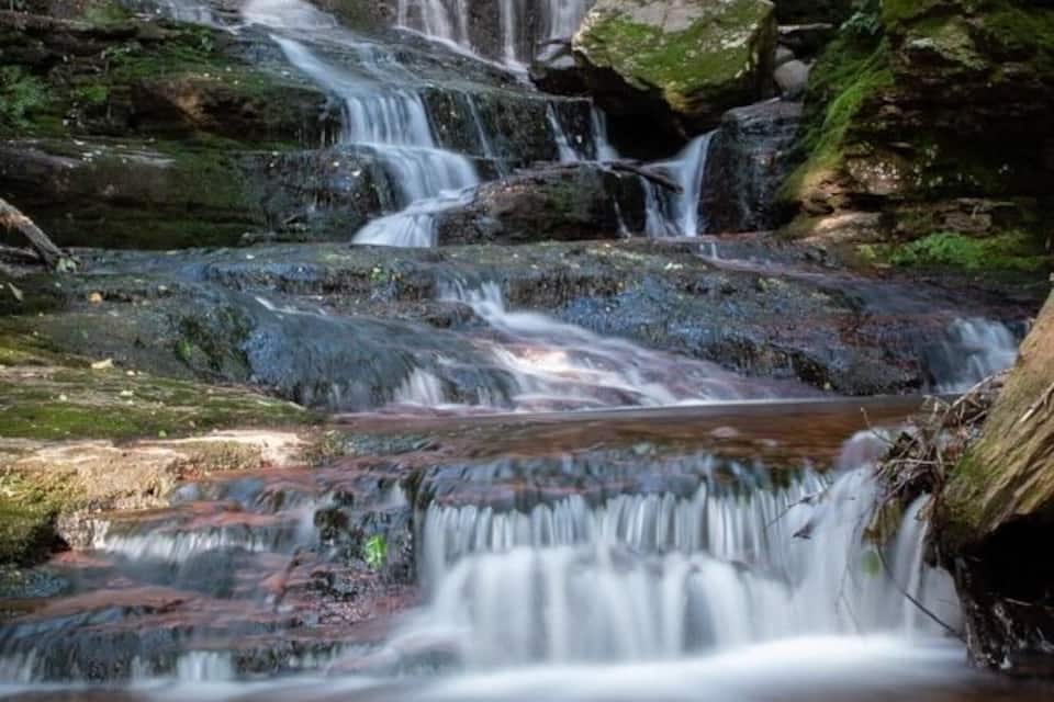 A multi-tiered waterfall in a forested area with moss-covered rocks and a calm pool at the base.