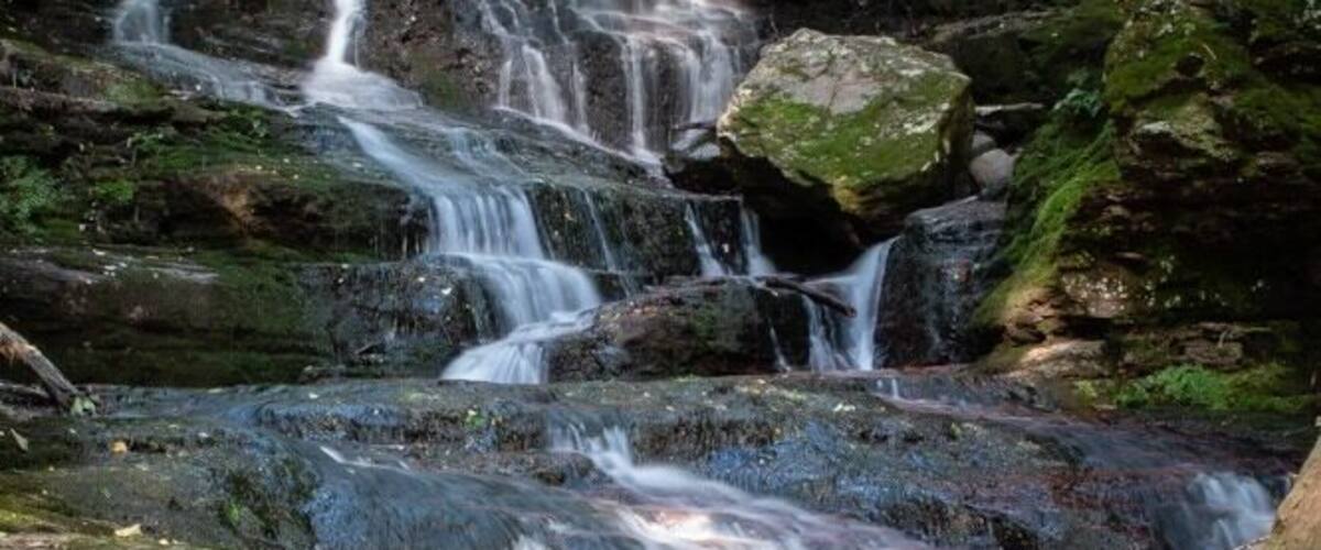A multi-tiered waterfall in a forested area with moss-covered rocks and a calm pool at the base.