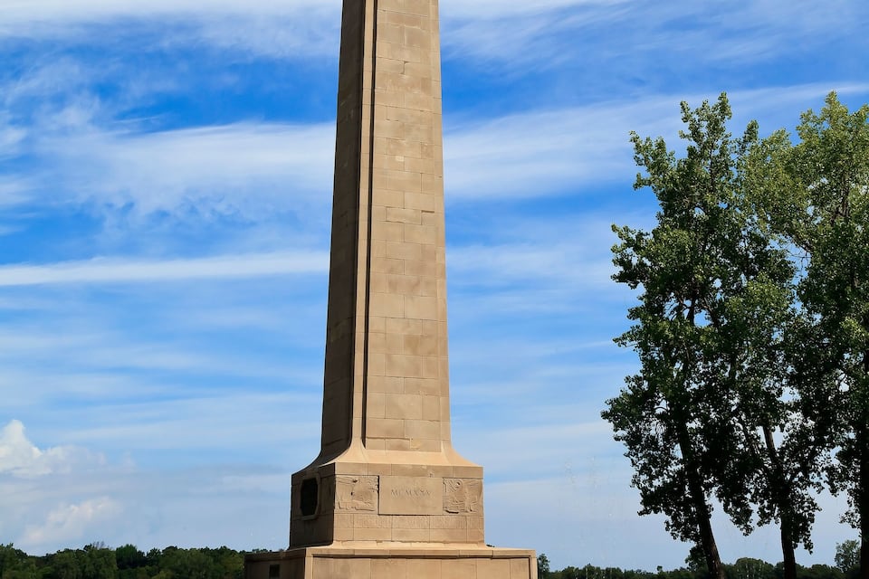 Um obelisco alto e bege, com o topo verde, erguido contra um céu azul com nuvens.