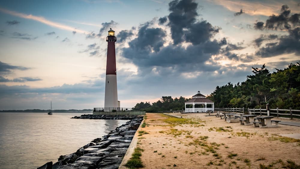 A lighthouse, a sandy beach, a wooden fence, and a gazebo by the water.